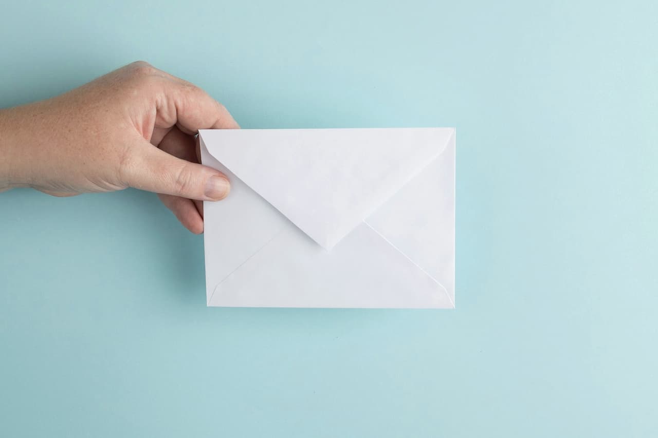A hand holding up a plain white envelope against a pale blue wall