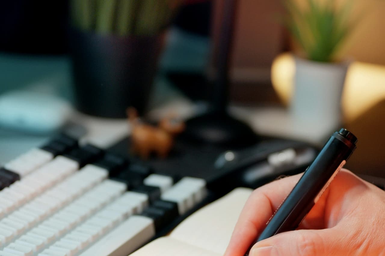 A hand writing in a notebook with checkboxes beside a keyboard