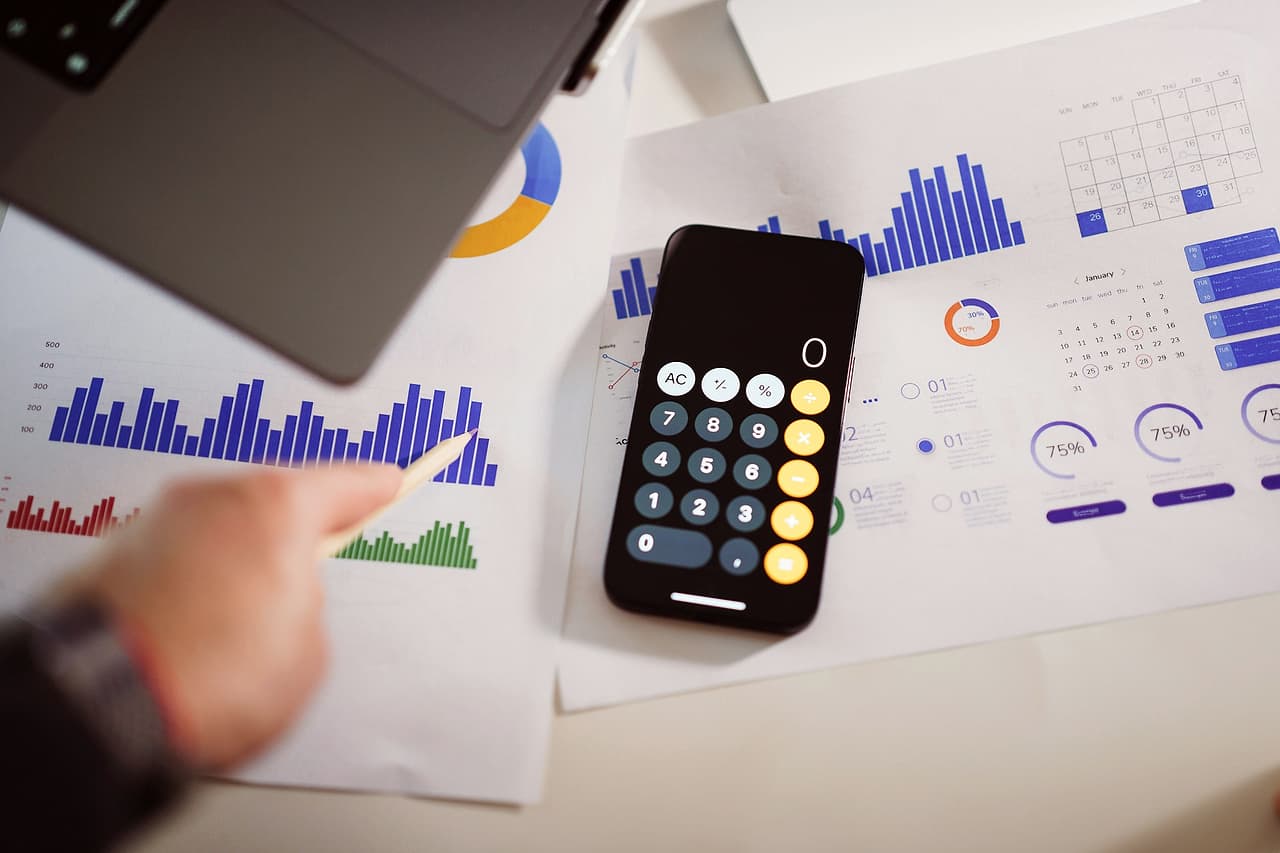 A phone calculator on a desk beside printed charts and a finger pointing at a graph
