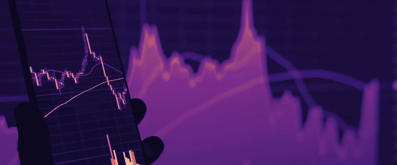 A hand holding a phone in front of a screen of price charts