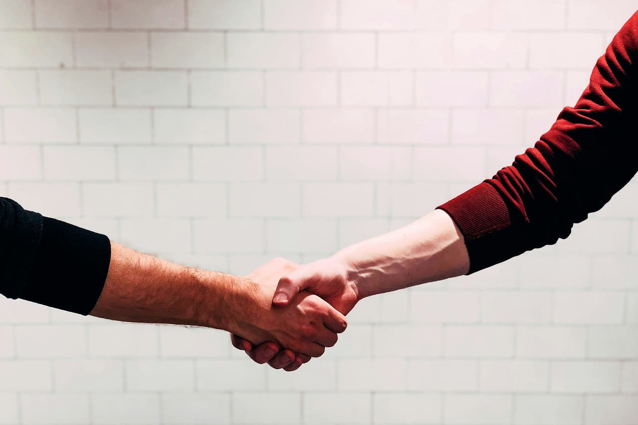 Two people shaking hands in front of a white tiled wall, faces out of frame