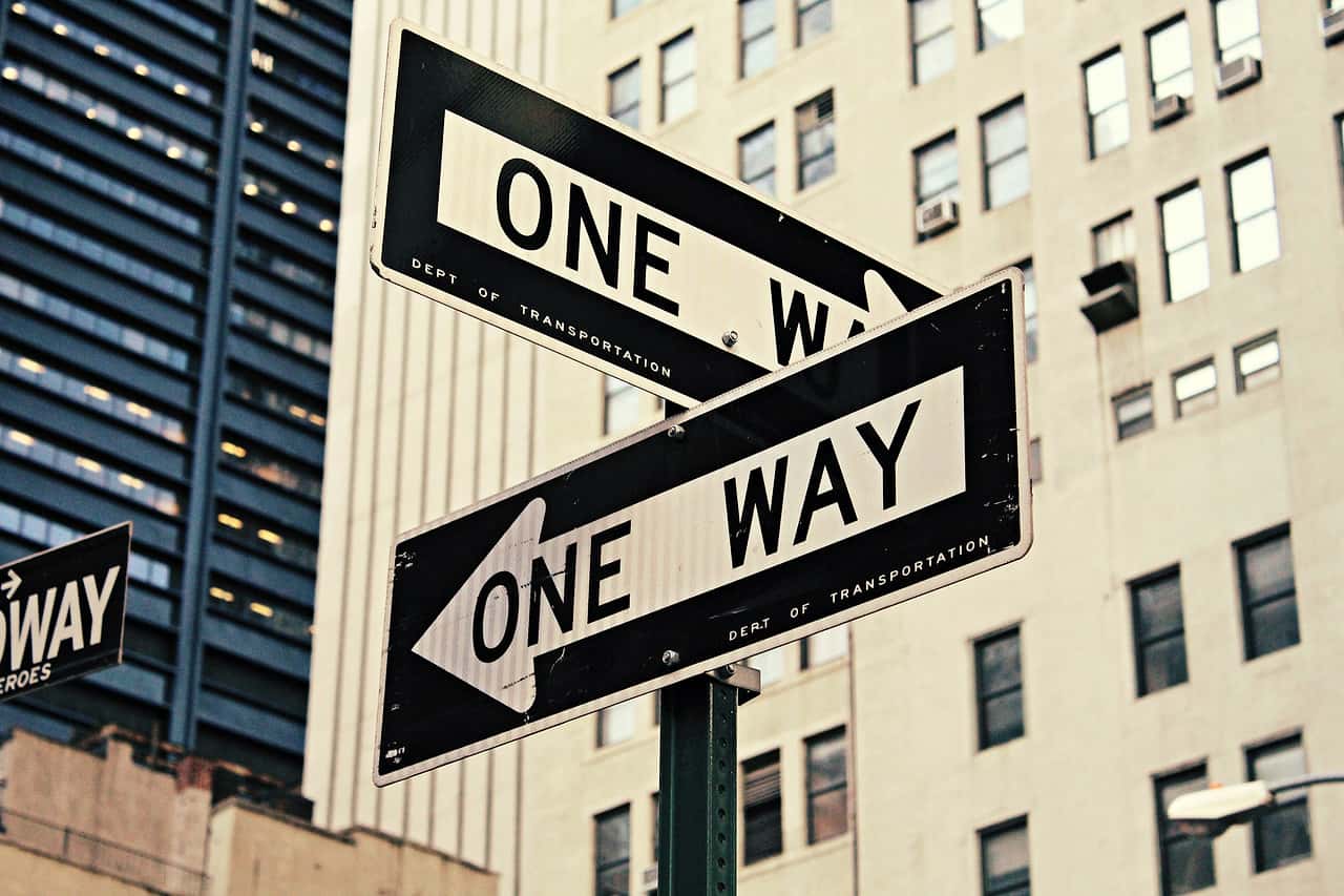 Two One Way street signs on a pole in front of city buildings