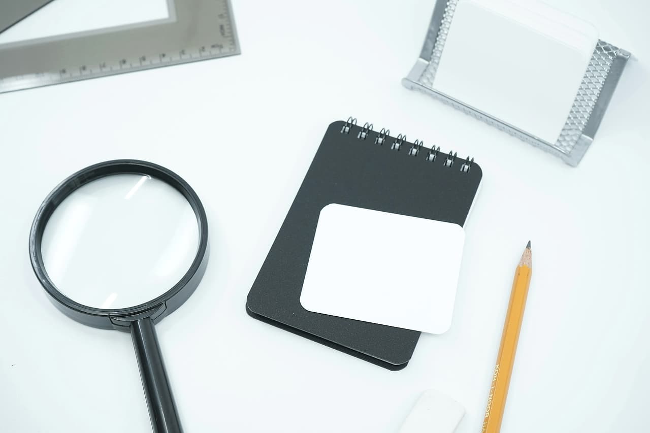A magnifying glass beside a notepad and pencil on a white desk